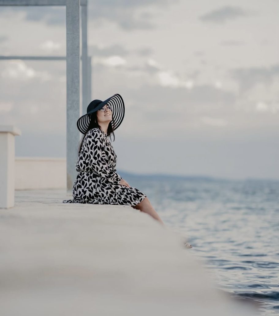 Frau in einem gepunkteten Kleid und breitem Hut sitzt am Ufer des Wassers.
