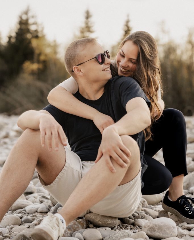 Junges Paar sitzt auf Kies, lächelnd und umarmend, mit Sonnenbrillen im Freien.