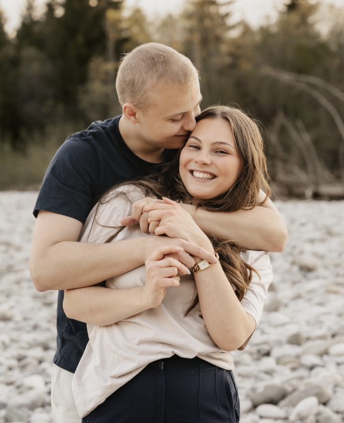 Paar umarmt sich lächelnd, im Hintergrund Kieselsteine und Bäume.