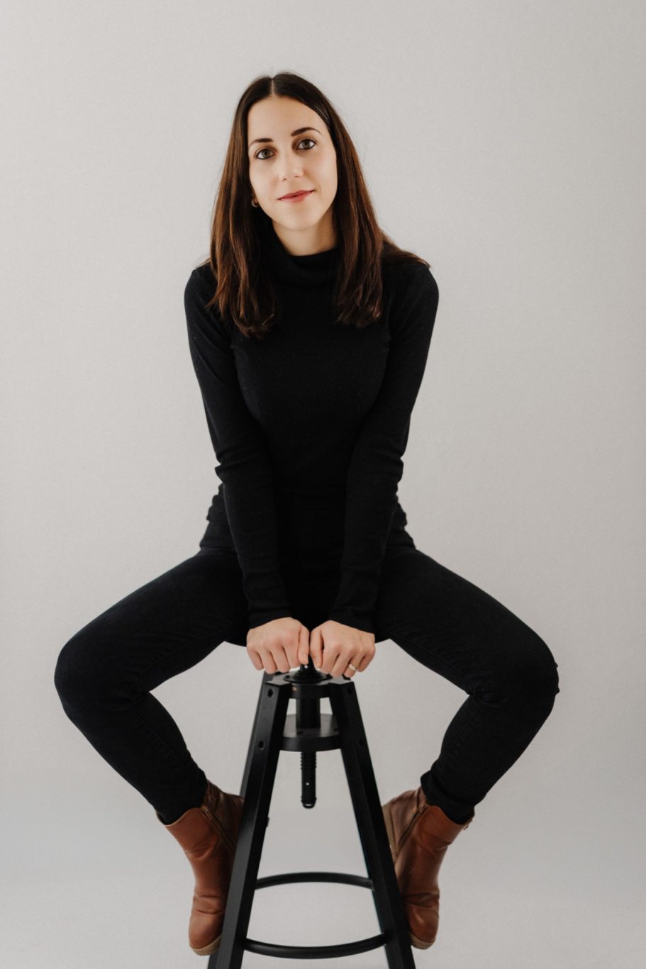 Eine Frau sitzt auf einem Hocker, trägt schwarze Kleidung und blickt direkt in die Kamera.