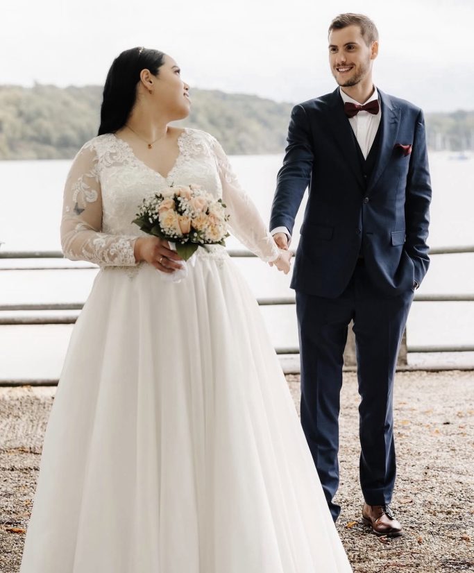 Braut und Bräutigam lächeln sich am Wasserufer an, umgeben von Natur.