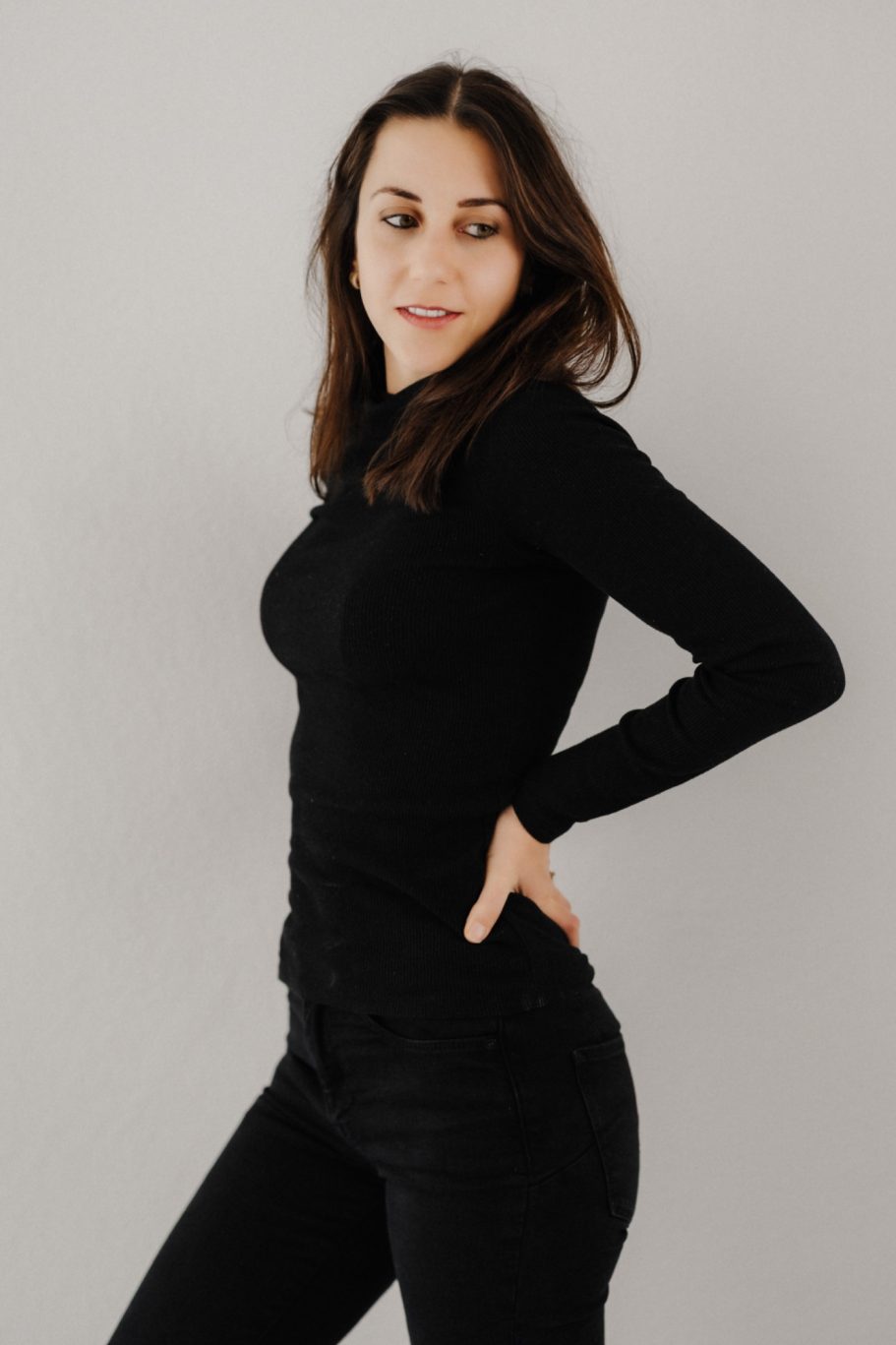 Frau in schwarzem Outfit, seitlich stehend, mit Hand an der Hüfte, lächelnd.