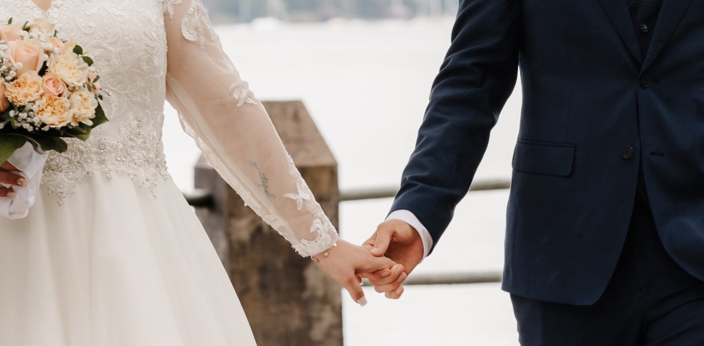 Ein Brautpaar hält Händchen, umgeben von einer romantischen Kulisse am Wasser.