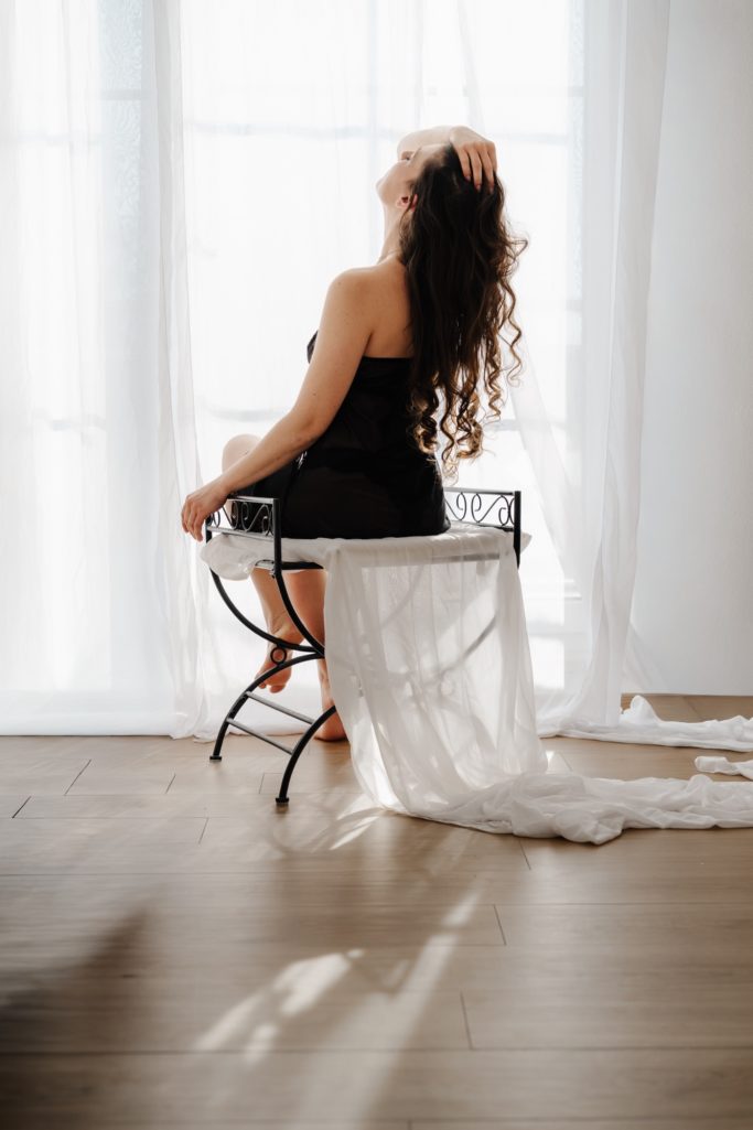 Frau mit Locken sitzt auf einem Stuhl vor einem Fenster, leicht zur Seite geneigt.