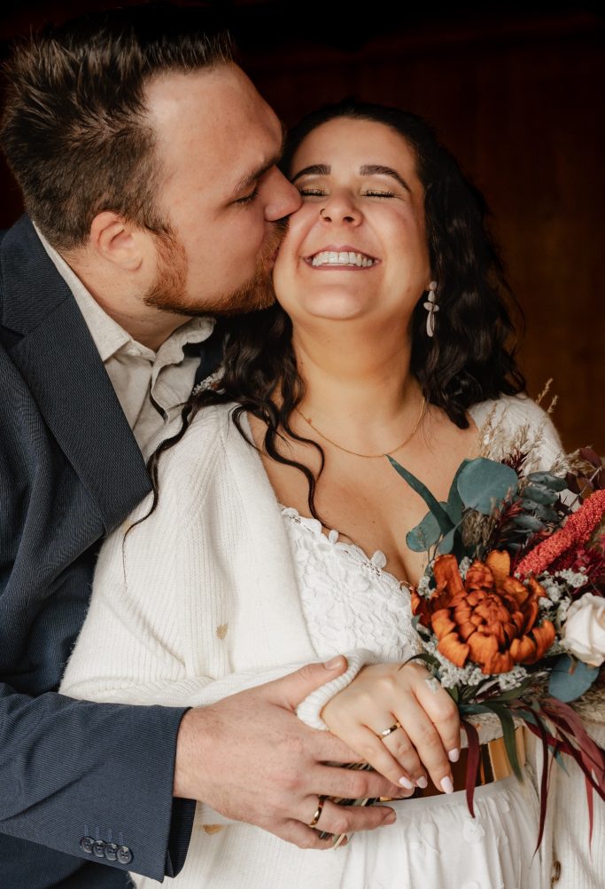 Ein lächelndes Brautpaar, der Bräutigam küsst die Braut, die einen bunten Blumenstrauß hält.