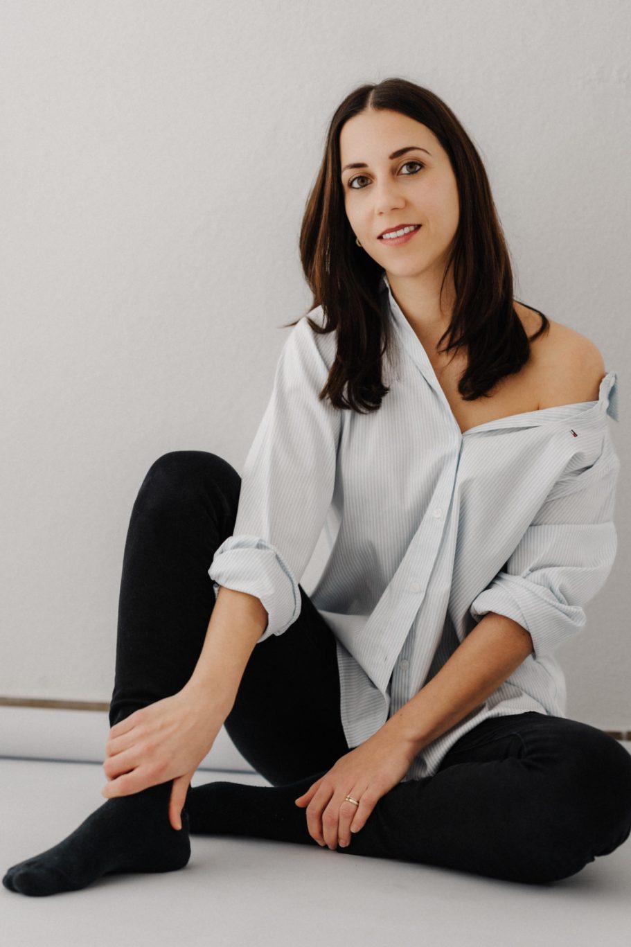 Junge Frau mit einem offenen Hemd, sitzt an einer Wand und lächelt.