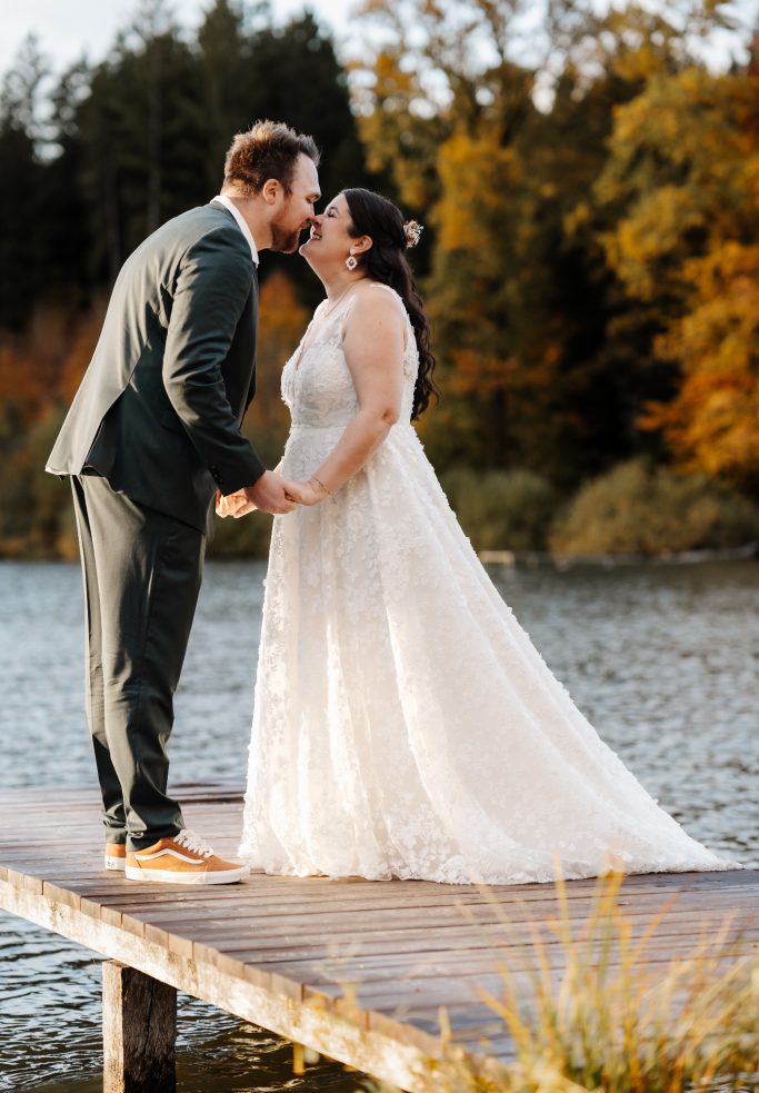 Braut und Bräutigam küssen sich auf einem Steg am Wasser, umgeben von herbstlichen Bäumen.