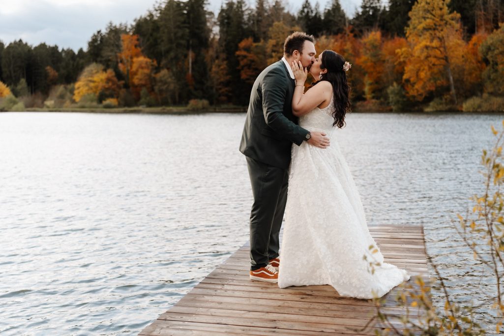 Ein Brautpaar küsst sich auf einem Steg am Wasser, umgeben von herbstlichen Bäumen.