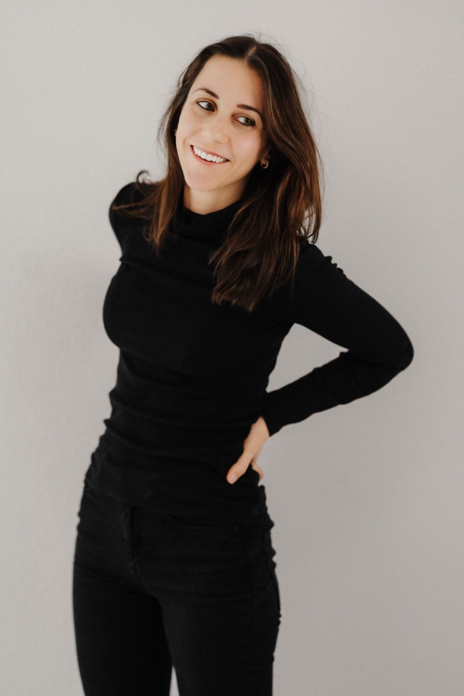 Eine lächelnde Frau in schwarzem Pullover und Jeans vor grauem Hintergrund.