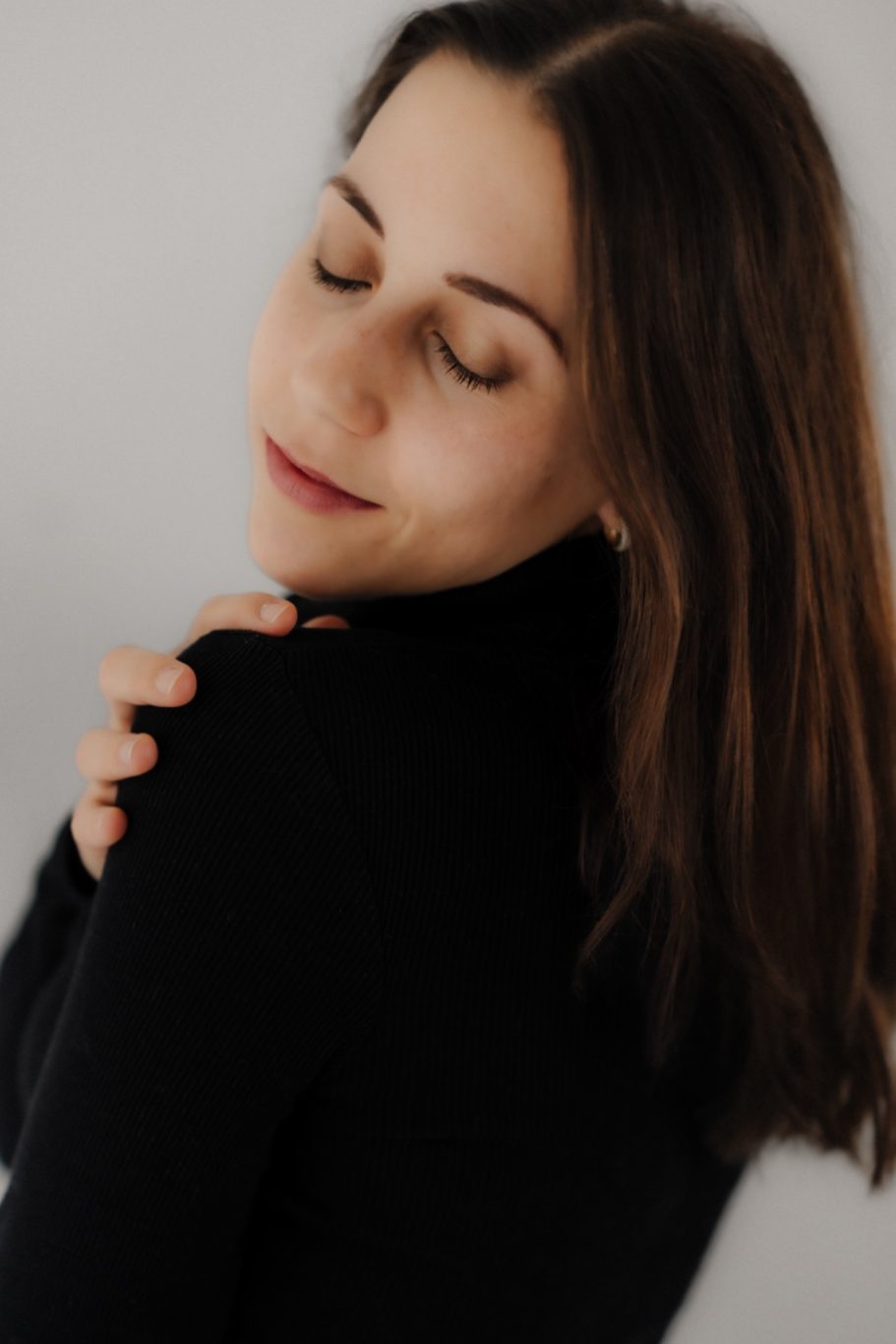 Frau mit geschlossenen Augen, die leicht lächelt und sich sanft an der Schulter berührt.