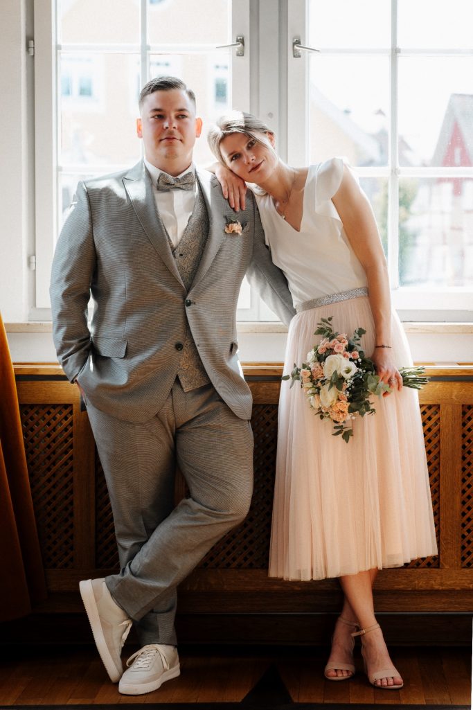 Ein Paar in eleganter Kleidung, lächelnd neben einem Fenster, mit einem Blumenstrauß.