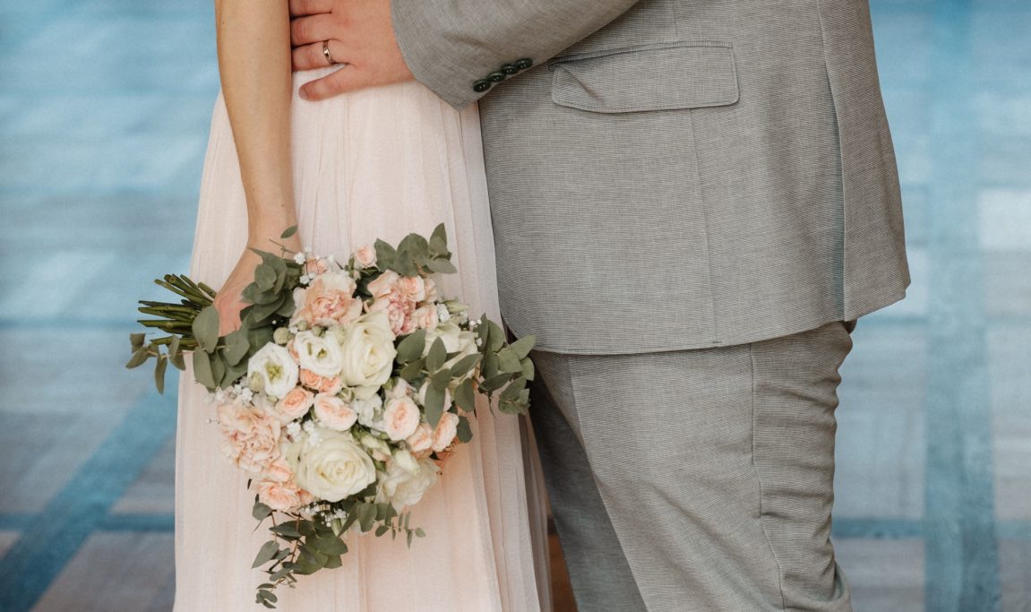 Ein Brautpaar steht zusammen, der Bräutigam in einem grauen Anzug und die Braut in einem rosa Kleid.