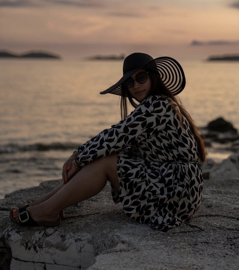 Frau in einem Tiermuster-Kleid und Sonnenhut sitzt am Ufer bei Sonnenuntergang.